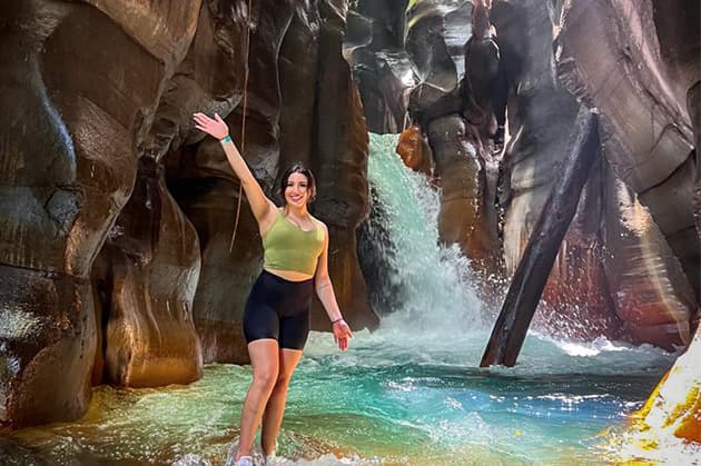 Standing in front of the breathtaking Cathedral Waterfall within a rocky canyon during the Cathedral Waterfall Expedition at Rincón de la Vieja, a person smiles and waves while wearing a green top and black shorts. The majestic rock formations and shimmering water create an awe-inspiring backdrop.