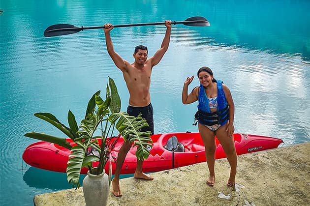 Two people cheerfully pose by their red kayak during the Cathedral Waterfall Expedition at Rincón de la Vieja; one holds a paddle overhead while the other waves. They're next to shimmering blue water, with a potted plant enhancing the scenic foreground.