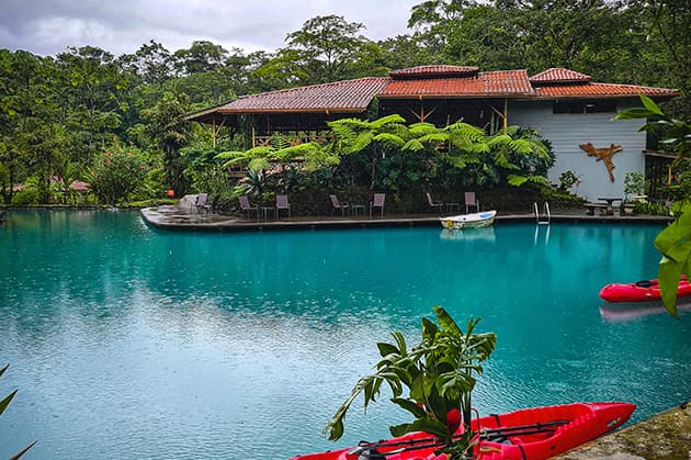 Located near Rincón de la Vieja, the Cathedral Waterfall Expedition offers a stunning tropical setting featuring a large blue pool surrounded by lush greenery. In the background, a building with a red-tiled roof stands prominently, and vibrant red kayaks in the foreground promise adventure.