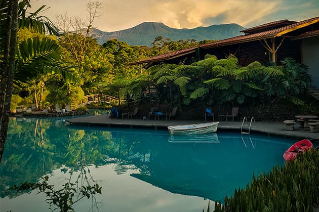 Experience the serene atmosphere of the Cathedral Waterfall Expedition at Rincón de la Vieja, featuring a tranquil setting where a boat glides effortlessly on pristine waters, encircled by vibrant foliage and breathtaking mountain vistas in the backdrop.