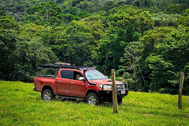 A red pickup truck is parked on a grassy field within the lush surroundings of Rincon de la Vieja, with a person visible inside.