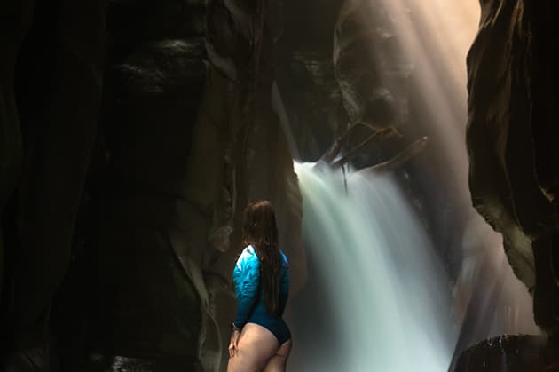 Standing amidst the rocky landscapes of a canyon during the Cathedral Waterfall Expedition at Rincón de la Vieja, a person gazes at a majestic waterfall as sunlight streams through.