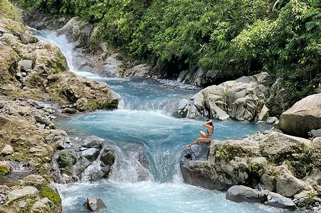 A person sits on a rock beside a clear blue river at Rincon de la Vieja, surrounded by lush greenery and rocky terrain.