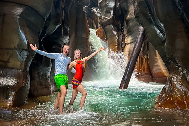 Two people stand in a rocky cavern at the Cathedral Waterfall Expedition in Rincón de la Vieja, with a waterfall cascading behind them. They pose cheerfully in their swimwear, surrounded by the clear, inviting water.