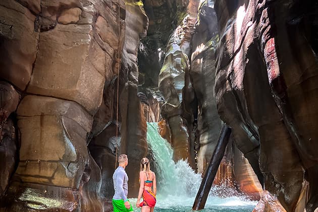 Two individuals dressed in swimwear are situated within the rocky canyon of Rincón de la Vieja, where the awe-inspiring Cathedral Waterfall Expedition serves as their stunning backdrop.