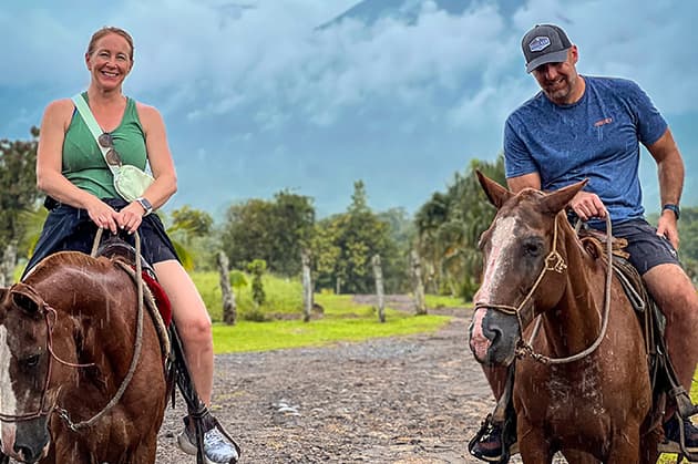 Two people riding horses on a gravel path with lush greenery and a cloudy sky in the background, evoking the tranquil beauty of Rincon de la Vieja.