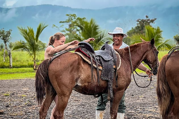 A person adjusts a saddle on a brown horse while another holds the reins, set against the stunning backdrop of Rincon de la Vieja's majestic palm trees and mountains.