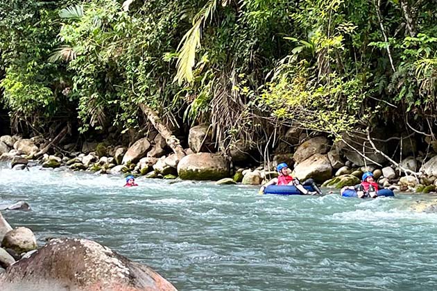 Participants wearing helmets and life jackets are enjoying the Tubing in Rincón de la Vieja experience, surrounded by lush green foliage and rocks as they float down the river.