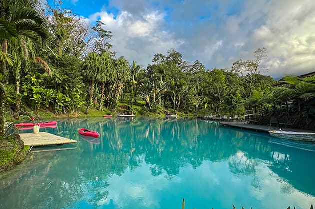 A serene pool set against the backdrop of Cathedral Waterfall Expedition in Rincón de la Vieja is embraced by verdant foliage and trees under a partly cloudy sky, with inflatable rafts and a small dock drifting gently on its surface.