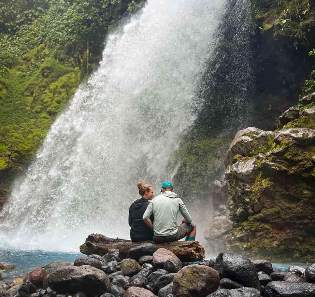 Two people sit on a rock by a waterfall at Rincon de la Vieja, enveloped in lush greenery and the soothing sound of flowing water.