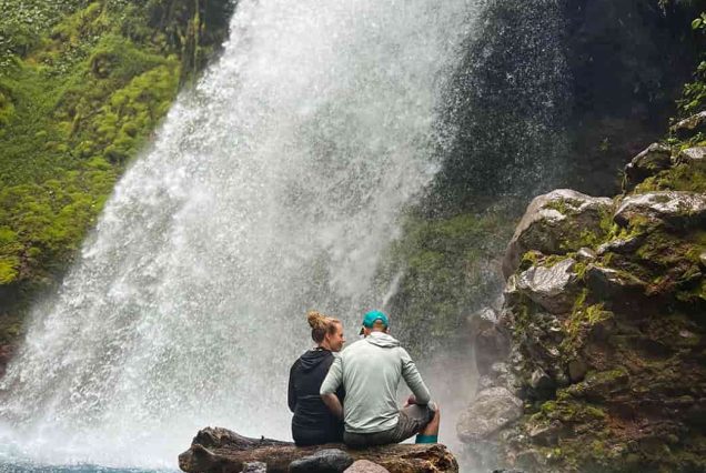 Two people sit on a rock by a waterfall at Rincon de la Vieja, enveloped in lush greenery and the soothing sound of flowing water.