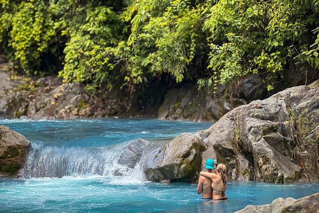 Two people swim in a natural turquoise pool at Rincon de la Vieja, surrounded by rocks and lush green foliage.