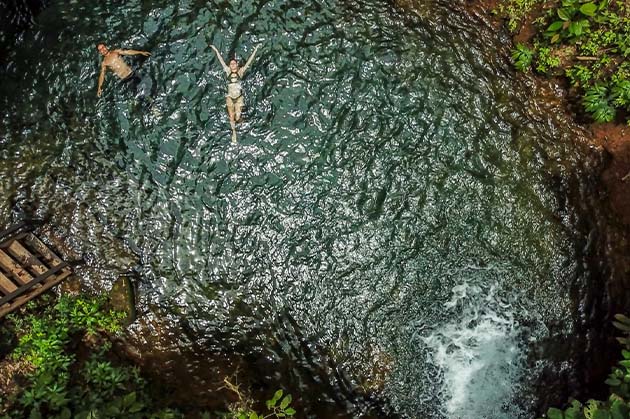 Two people float in a small, circular pool surrounded by the lush greenery typical of Rincon de la Vieja. They lie on their backs in the water, basking in tranquility. Steps can be seen on the left side, inviting a gentle descent into this serene oasis.