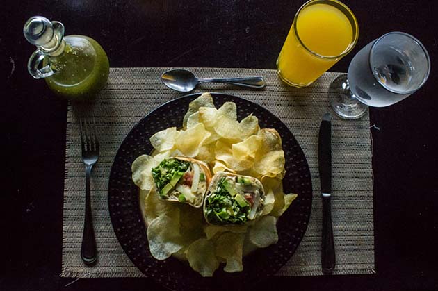 A plate of wraps and potato chips sits on a woven mat, paired with a refreshing glass of orange juice and water. Nearby, a bottle of green dressing awaits, reminiscent of the vibrant flavors found near Rincon de la Vieja.