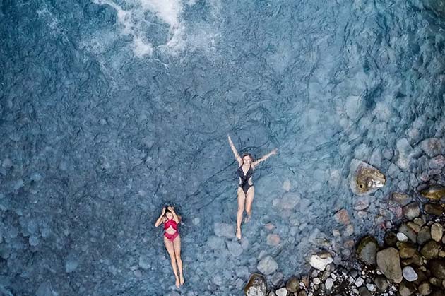 Two people float in the clear blue waters near the rocky shorelines of Rincon de la Vieja, wearing swimsuits.