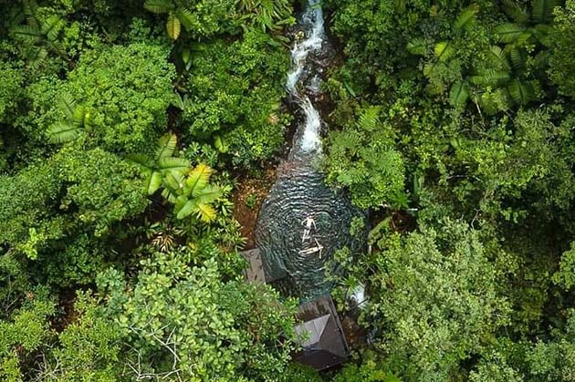 Aerial view of a lush green forest in Rincon de la Vieja, with a small river flowing through it. A person floats serenely in the water beside a quaint wooden structure.