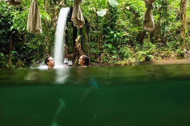 Two people bask in a warm pool under a waterfall, surrounded by the lush greenery of Rincon de la Vieja. Towels hang on a bar above the water, adding to the tranquil setting.