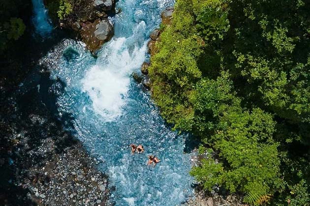 Aerial view of two people swimming in a blue river surrounded by lush green foliage and rocks, capturing the serene beauty of Rincon de la Vieja.